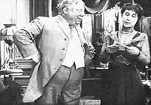 A black-and-white film still of an overweight man, hands on hips, staring at a woman to the right.