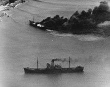Black and white aerial photo of two ships near a coastline. One of the ships is on fire.