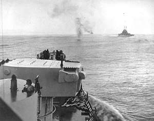 A plume of smoke rises from a crashed aircraft ahead of a naval warship