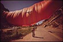 An enormous volume of fabric hangs from a wire across a valley. I the foreground is a telephone pole and several people looking up.