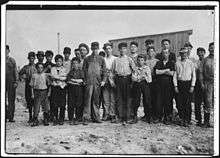 20–25 males facing camera in a row, mostly children. Ages vary. They are in work clothes. Some are dirty. Some wear caps. They stand on dirt with a wooden building behind them.