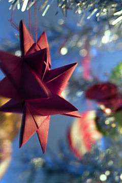 An out-of-focus close-up of a part of a Christmas tree with glitter garlands and blurry decoration. In-focus in the left half of the picture, suspended from one of the twigs, is a red cardboard-woven Froebel star. Four tips and seven prongs are visible.