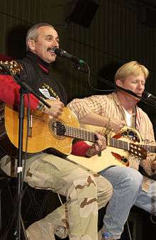 A grey-haired man with a moustache playing the guitar and singing into a microphone.  A blond-haired man, also playing a guitar, is in the background.