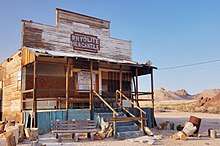 A small, wooden one-story building with a false second-story front rests on a flat expanse of sparsely-vegetated gravel near some reddish rocks and low hills under a cloudless light-blue sky. A dozen or so sawed logs, a rusting barrel, and a bench for sitting, have been placed here and there in front of the building. A sign on the false front says, "Rhyolite Mercantile".