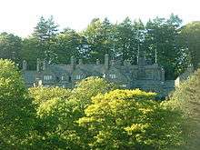 Peeping over the tops of trees in the foreground is a stone house with five gables of different sizes.  More trees are in the background.