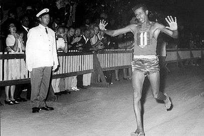 Abebe Bikila crossing the finish line, barefoot with hands raised, an Italian official in the background with a crowd of spectators behind a fence
