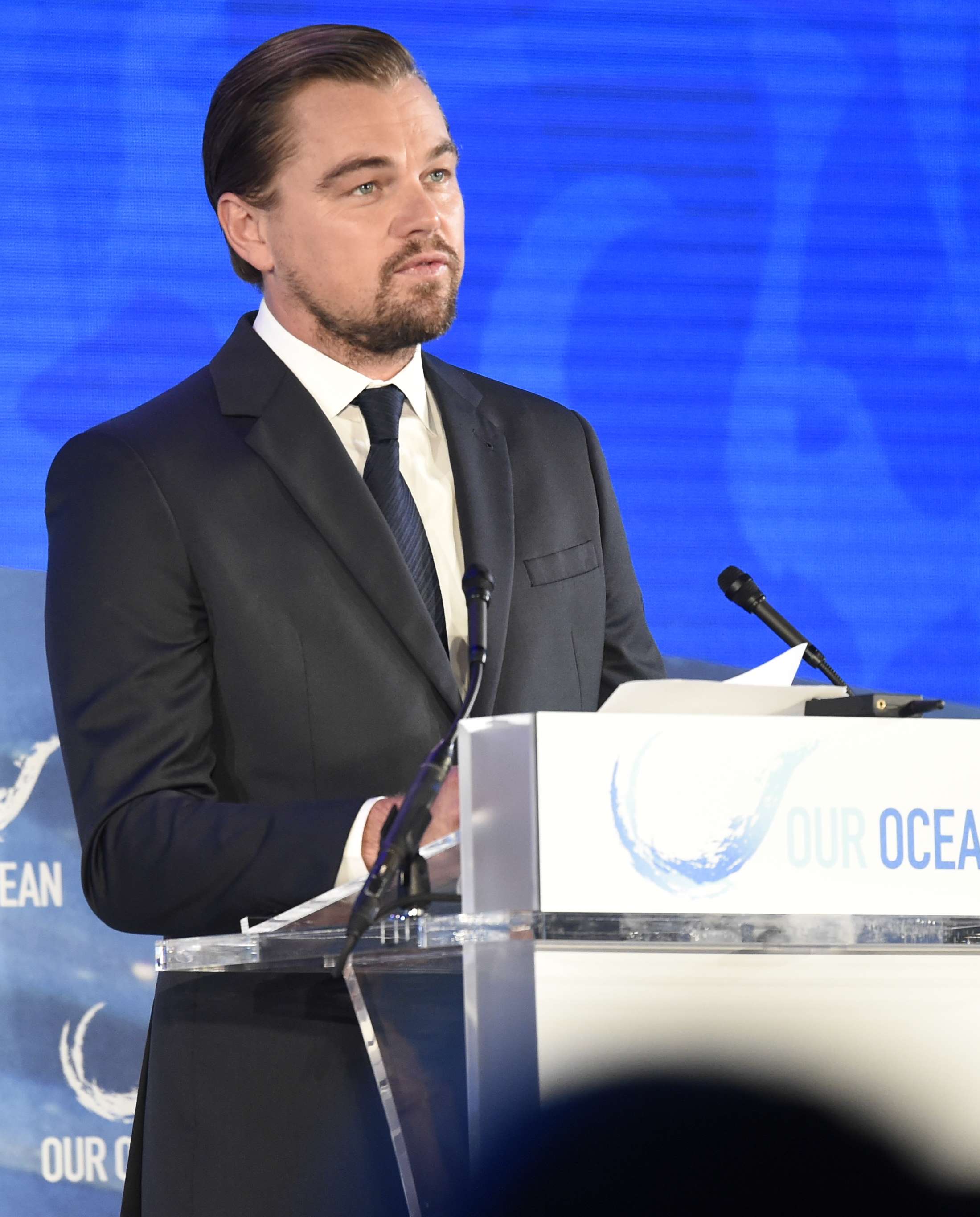 A white man with brown hair, combed back, and a trim mustache and beard wearing a dark jacket and tie stands before a lectern speaking in front of a blue background
