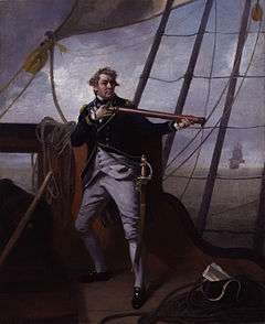 A large man in a blue uniform strikes a dramatic pose holding a telescope on the quarterdeck of a ship. In the distance another ship can be seen with its sails set.