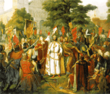 A bishop blesses a crowned man while people, many of them on their knees, whatch them