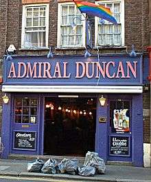 A blue/purple public house with large prominent signage as "The Admiral Duncan". A rainbow flag is above it, and a number of garbage bags are on the pavement in the foreground.