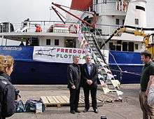 Two men in suits in front of a ship. The vessel is titled the Rachel Corrie and a banner reads "Freedom Flotilla"