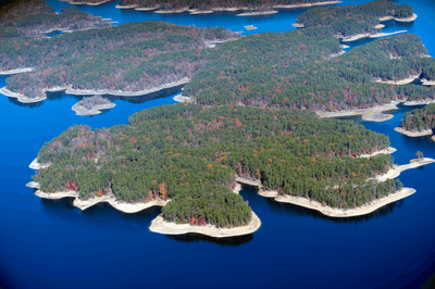 Aerial view of a calm, shiny blue lake and matriculating around wooded peninsulas covered in green, orange and red foliage