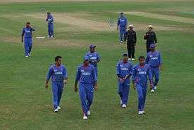 Nine men in a field wearing a blue outfit. Two men wearing a black outfit, along with a cricket pitch in the background, can be seen.