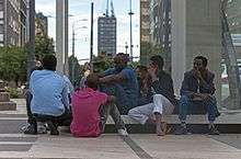 A group of black African men sitting or squatting on a low bench next to a glass wall in a large city square. In the rear can be seen a street with a tall rectilinear skyscraper