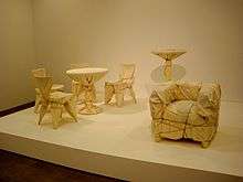Four cafe chairs and a table, a high table, and an upholstered chair are wrapped in fabric and rope.