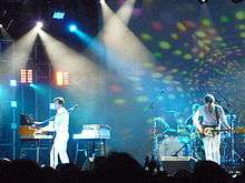 Three men play instruments on a brightly lit stage. Two of the men wear white clothing; they play a guitar and a keyboard respectively. The third man wears a black shirt and sits behind a drum set, playing.