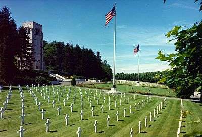 Photograph of field of graves marked with white crosses, with two American flags and a stone memorial building