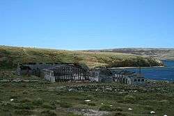 A cluster of ruined industrial buildings sits on the shore of a green, treeless landscape.