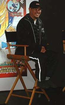 Akira Toriyama sitting on a chair smiling wearing black clothing with glasses and chin facial hair.