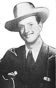A man wearing a white cowboy hat and dark jacket, smiling broadly and holding a guitar