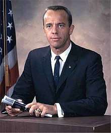Shepard sits at a table wearing a dark suit and holding a microphone. He is wearing his Naval Academy class ring, and an astronaut pin on his lapel. In the background is an American flag