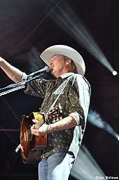 A fair-haired man wearing a white cowboy hat, a green shirt and blue jeans, playing a guitar and singing into a microphone