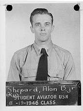Shepard, in Navy uniform short and tie, stands before a blackboard on which is stencilled "Student aviator USN – 8-19-1946 class. Above that is written in chalk: "Lt (jg) Shepard, Alan B. Jr"
