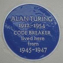 A Blue plaque on a white wall with the words "Alan Turing 1912–1954 CODE BREAKER lived here from 1945&nbsp;– 1947
