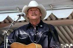 A fair-haired man wearing a white cowboy hat, singing into a microphone and playing a guitar