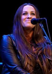 An image of a woman with long brown hair and a leather jacket. She is looking toward the camera while performing on stage.