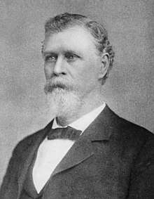 A man with gray hair, beard, and mustache wearing a black jacket and neck tie and a white shirt