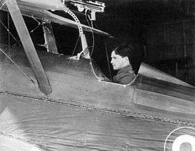 Side view of dark-haired man in open cockpit of biplane equipped with machine-gun on upper wing