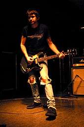 A male musician, Steve Albini, with an electric guitar strapped over his shoulders in a playing position. He is wearing blue jeans with large rips in both knees.
