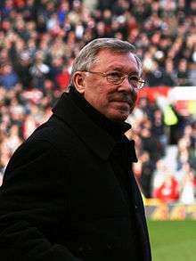Sir Alex Ferguson appears in the image. He is an older man wearing glasses and a black coat.