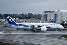 Boeing 787 in launch customer All Nippon Airways' blue and white livery. In the background are two assembly halls, with huge doors facing left. Vehicles are parked in front of the halls.
