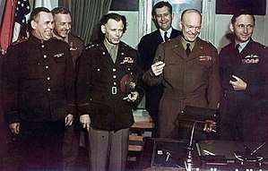 Group shot of six smiling men (and one barely visible woman), each in a different military uniform, standing behind a writing desk. Eisenhower is holding three fountain pens. In the background are flags, including the US flag.