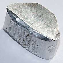 A silvery white steam-iron shaped lump with semi-circular striations along the width of its top surface and rough furrows in the middle portion of its left edge.