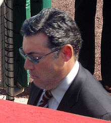 A dark-haired man wearing sunglasses, a dark jacket, and a red-and-white striped tie