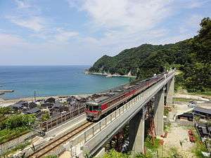 Diesel multiple unit train crossing the concrete Amarube Viaduct. A green hillside is visible on the right, and a bay of the Sea of Japan on the left.