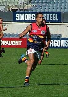 A male athlete with dark hair wearing a sleeveless jersey and shorts runs on the grass surface of the playing arena.