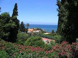 Varied foliage and flowers in the foreground the landscape slopes downward toward a tennis court and buildings lying near the coast