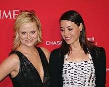 Amy Poehler and Aubrey Plaza stand side by side in front of a red background. Plaza appears to be looking at Poehler's cleavage.