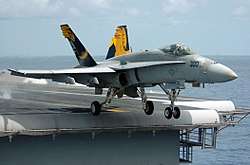 F/A-18 Hornet fighter departing aircraft carrier. A gray-overall aircraft, with blue and yellow fins, has just left the edge of carrier's runway, as evident through the extended landing gear.