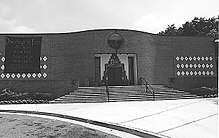 A black and white picture of a one-story brick building. The entrance door is in the center of the building and three rows of white diamond shaped tiles decorate either side.