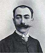 Head and shoulders photo of a youngish man with receding dark hair and large moustache, wearing striped suit