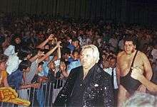 Bobby Heenan (front), a Caucasian man in a black sequin jacket, leads André the Giant to the ring