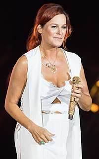 A red-haired woman performing on stage. She is wearing a white outfit and holding a golden microphone.