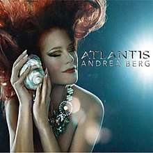 A red-haired woman underwater with her eyes closed. She is wearing a shiny pearl necklace. She is holding a shellfish against her cheek. The words "Andrea Berg Atlantis" are written in silver on the right.