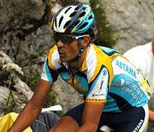 A cyclist in a blue and yellow jersey with white trim. His bicycle is not visible, but he is in riding position. He wears sunglasses, a helmet to match his jersey, and a bandage on his nose.