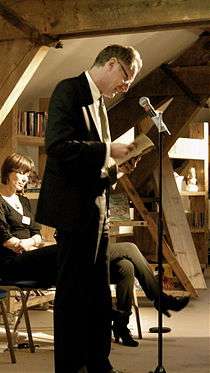 A tall man wearing a suit and holding a sheaf of papers stands, slightly stooping, in front of a microphone. A seated woman, and the crossed legs of another, are visible behind him.
